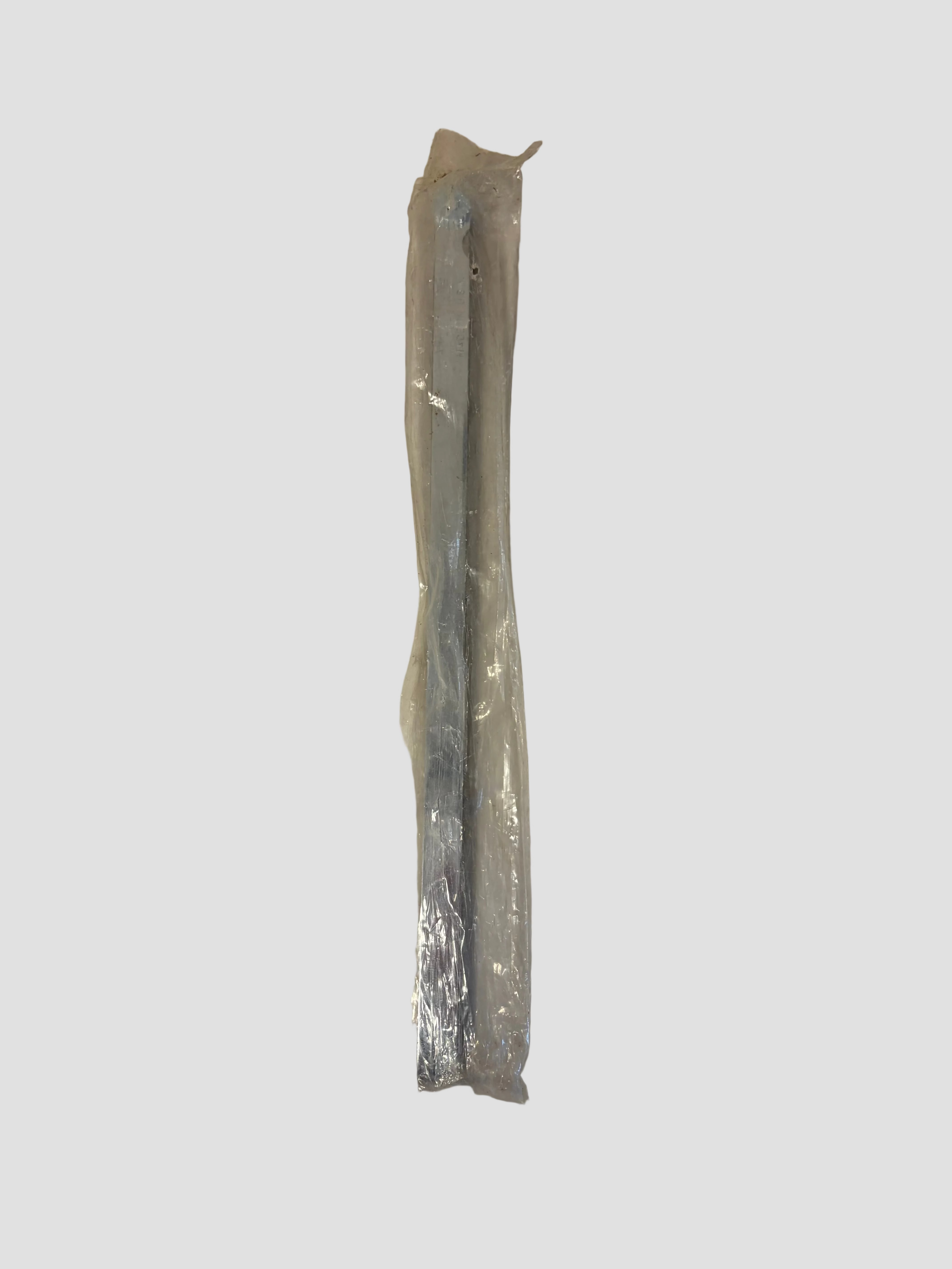 A long, thin metallic tool or rod sealed in clear plastic packaging, placed on a plain off-white surface.