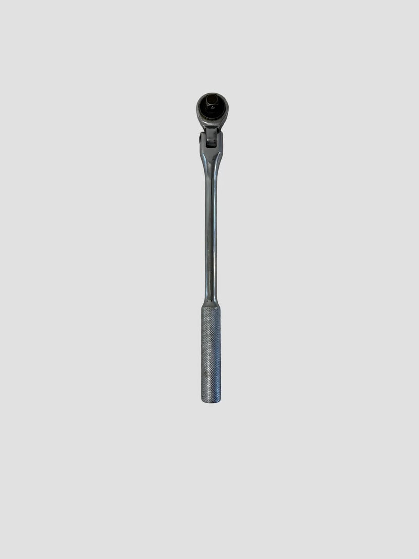 A chrome ratchet wrench placed on a light-colored surface. The wrench has a long, polished handle with a textured grip at the end for better control. Its ratcheting head features a square drive socket connector and a directional switch, showing a reflective metallic finish typical of durable hand tools used in mechanical work.