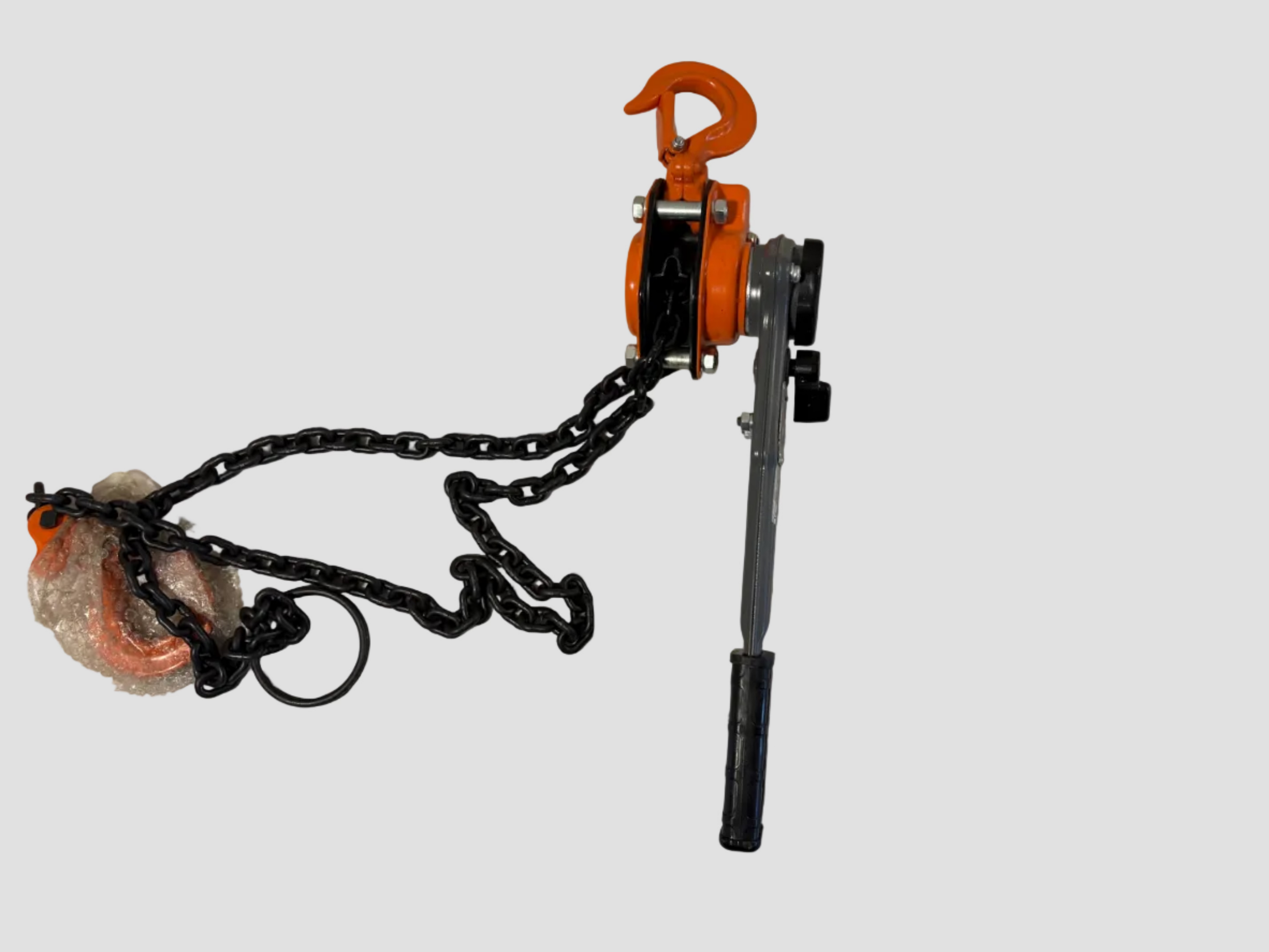 A manual chain hoist with a black metal chain, orange pulley housing and hook, and a gray handle, laid out on a plain off-white surface.