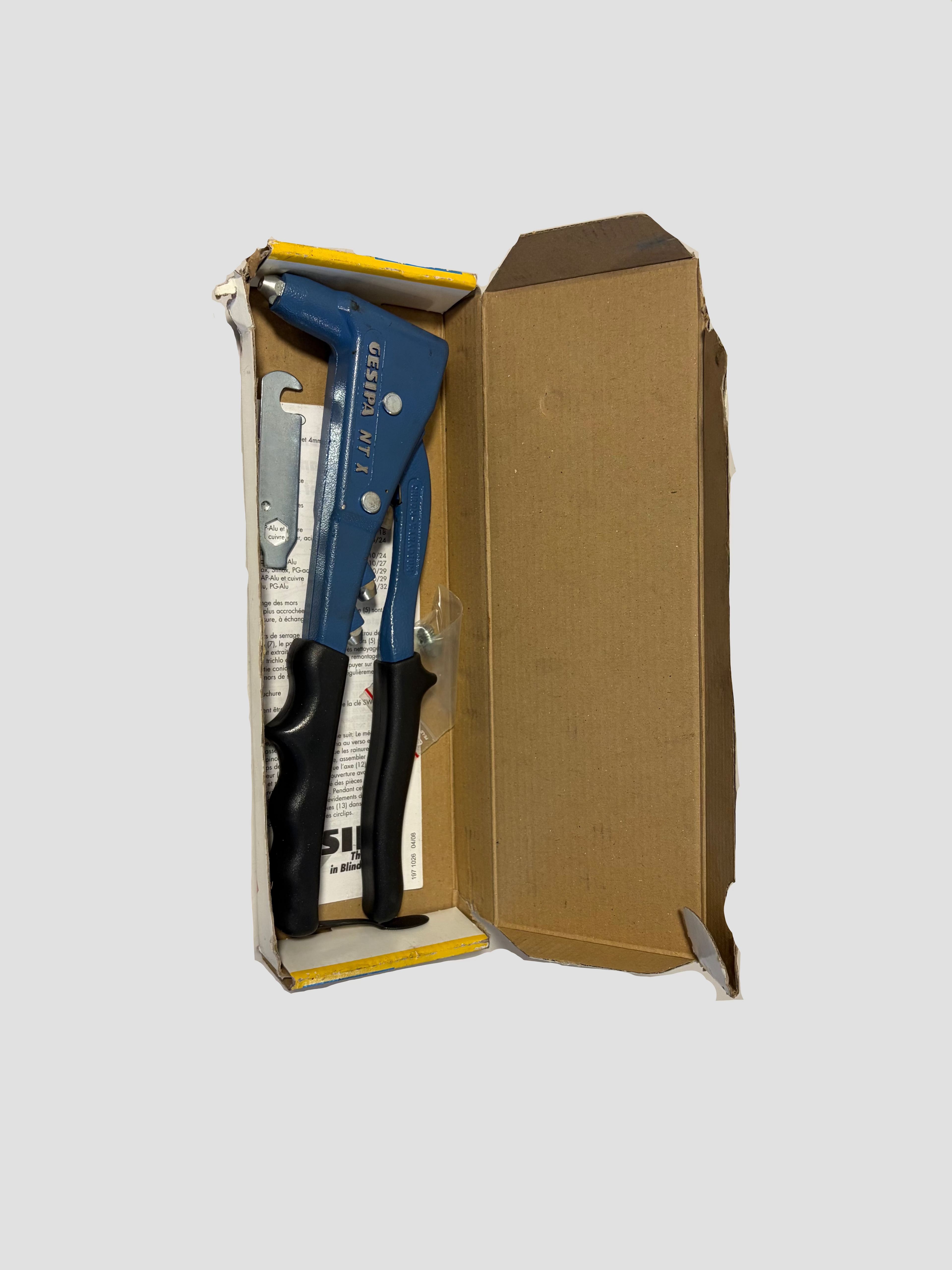 A blue and black hand tool with a metal spring-loaded clip is shown inside an open cardboard box with yellow trim, photographed against a plain off-white wall.
