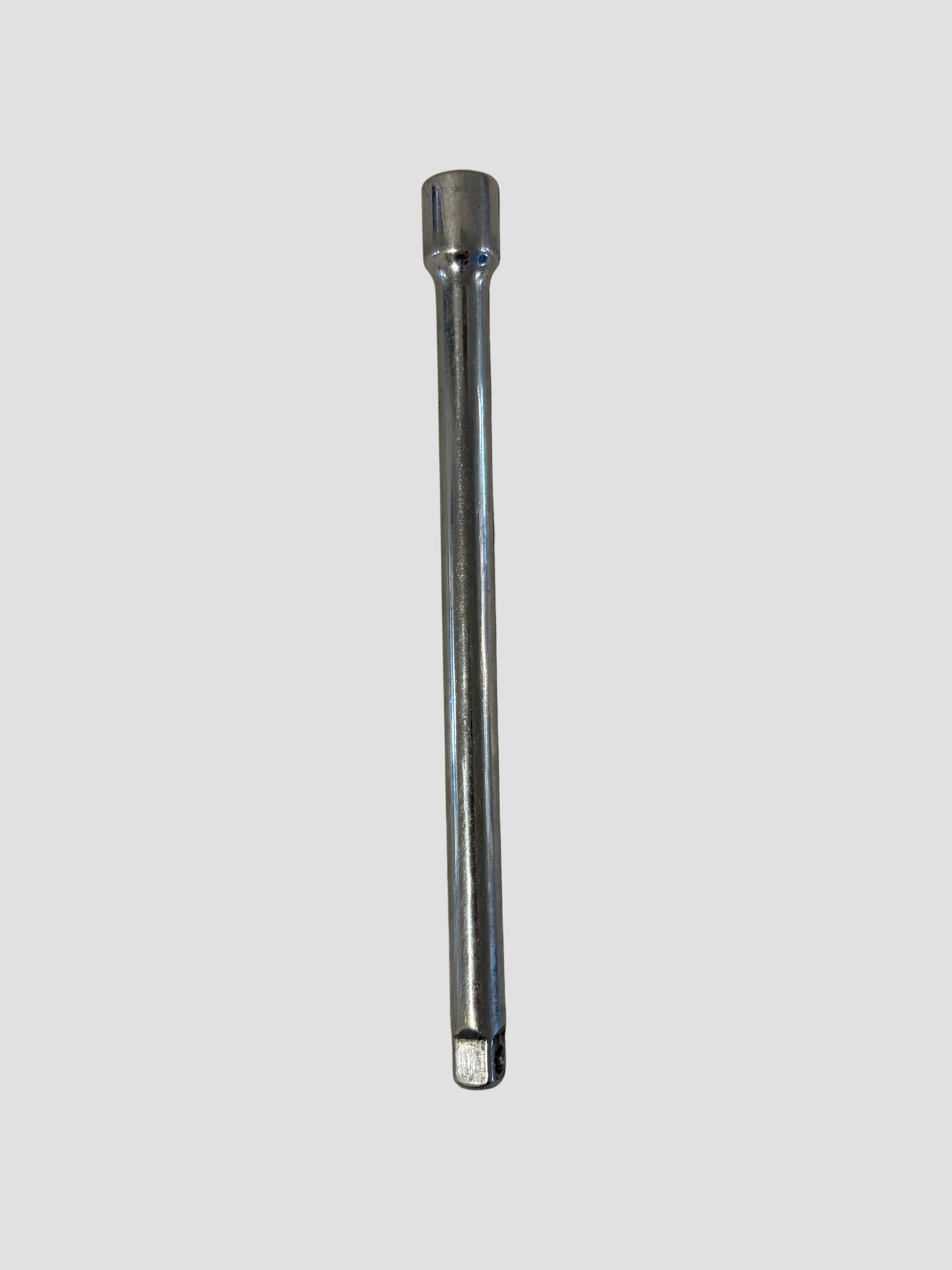 A chrome socket wrench extension bar lying flat on a light-colored surface. The tool has a smooth, cylindrical body with a square drive end on one side and a socket connector on the other. Its polished metallic finish reflects light, highlighting the durable steel construction and simple, functional design.