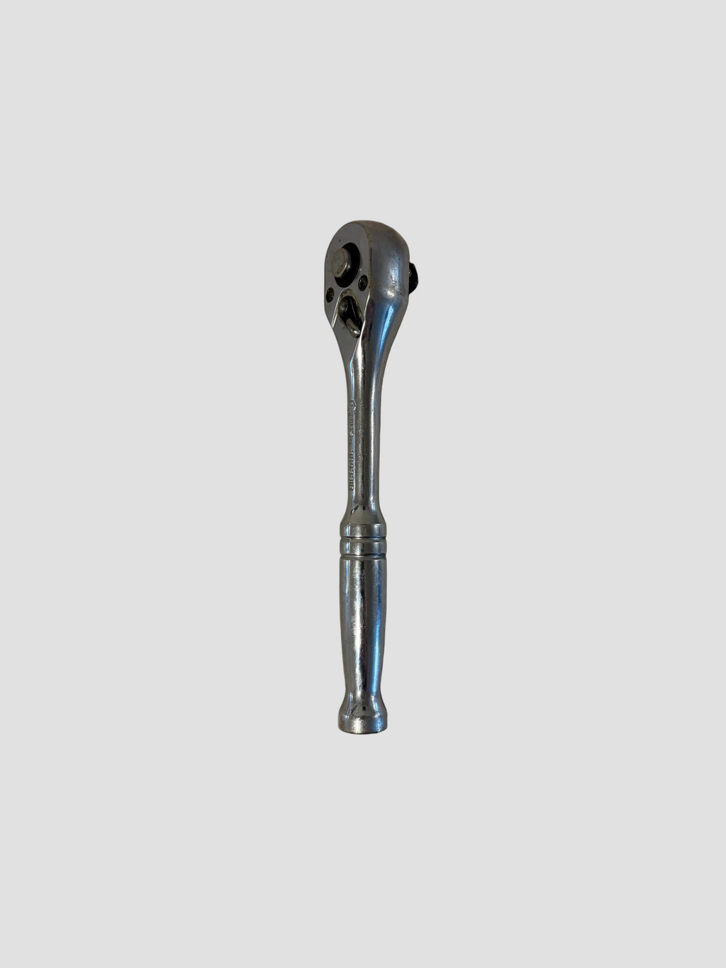 A chrome ratchet wrench lying flat on a light-colored surface. The wrench has a polished metallic finish with a short handle and a rounded ratcheting head featuring a square drive socket connector and directional switch. The reflective surface highlights its smooth contours and durable construction, typical of professional hand tools.