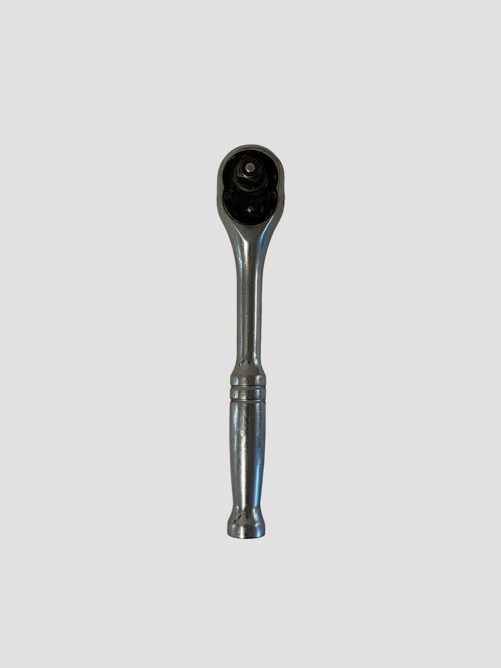 A chrome ratchet wrench lying flat on a light-colored surface. The wrench has a polished metallic finish with a rounded handle featuring two decorative grooves near the middle. Its ratcheting head includes a circular gear mechanism and a square drive socket connector, showing slight wear from use. The reflective surface highlights the tool’s smooth contours and durable construction.