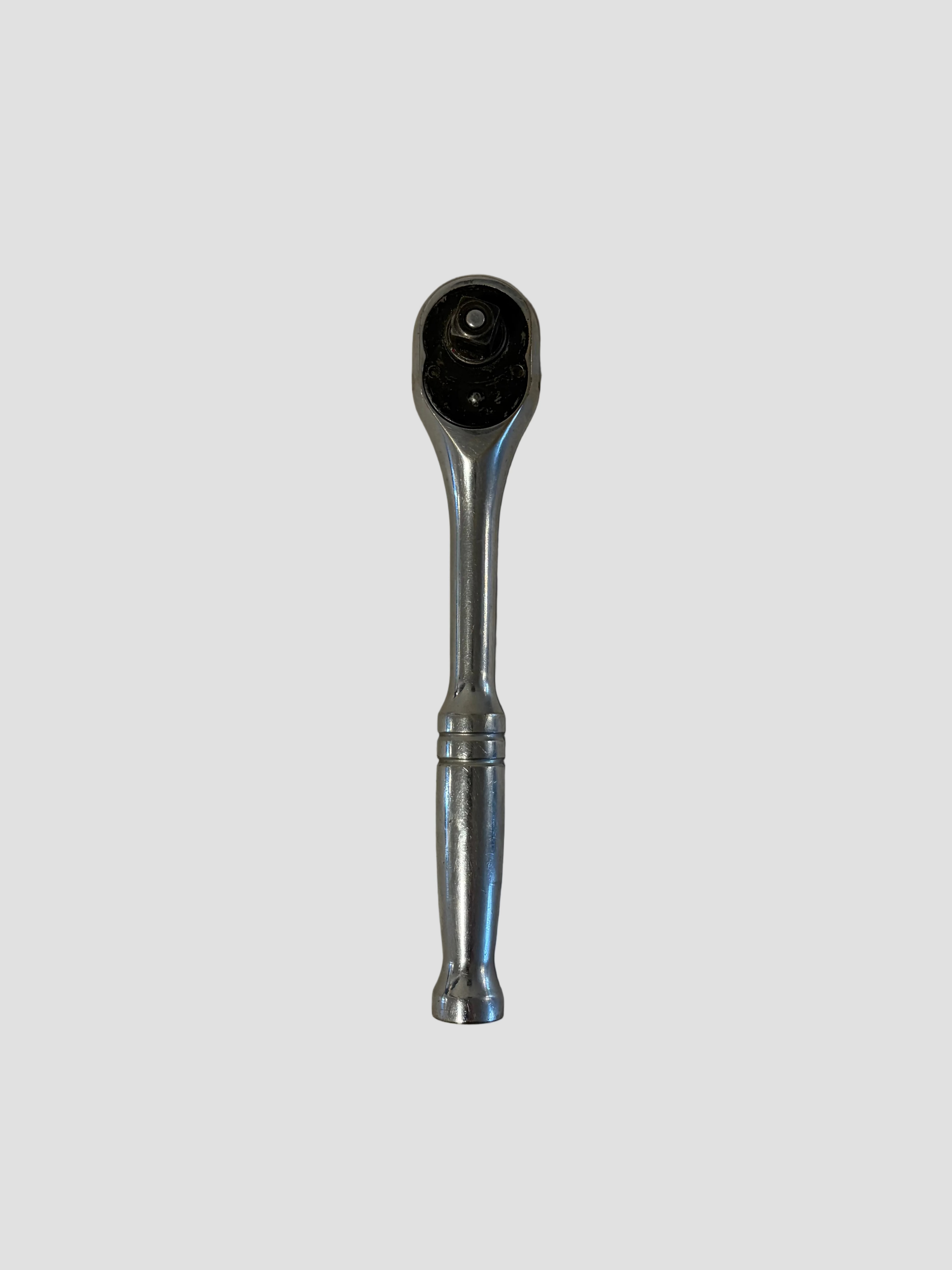 A chrome ratchet wrench lying flat on a light-colored surface. The wrench has a polished metallic finish with a rounded handle featuring two decorative grooves near the middle. Its ratcheting head includes a circular gear mechanism and a square drive socket connector, showing slight wear from use. The reflective surface highlights the tool’s smooth contours and durable construction.