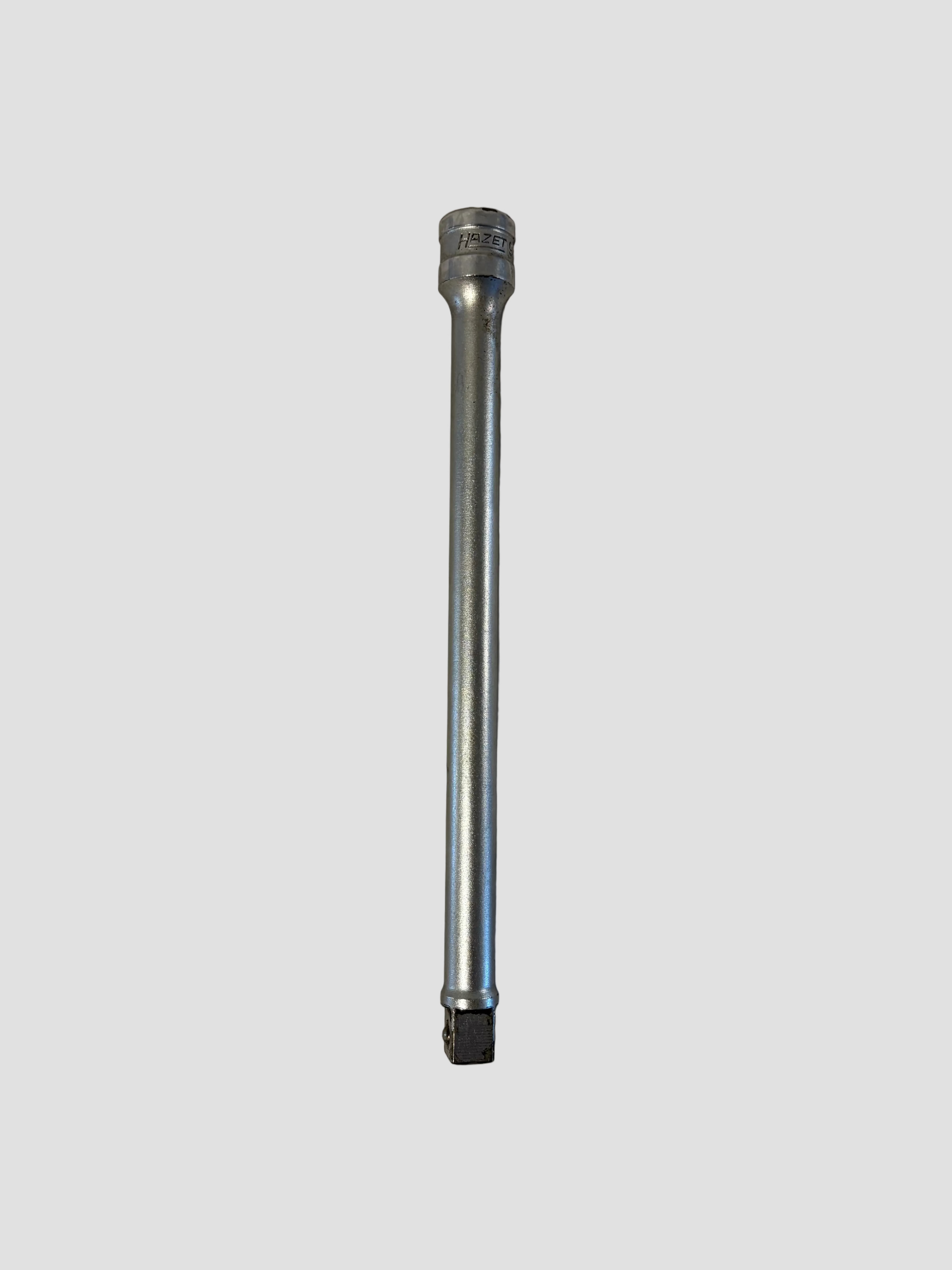 A chrome Hazet socket extension bar lying flat on a light-colored surface. The tool has a smooth cylindrical body with a square drive end at the bottom and a socket connector at the top engraved with the brand name “HAZET.” Its polished metallic finish reflects light, highlighting the durable steel construction and precision engineering typical of professional-grade tools.