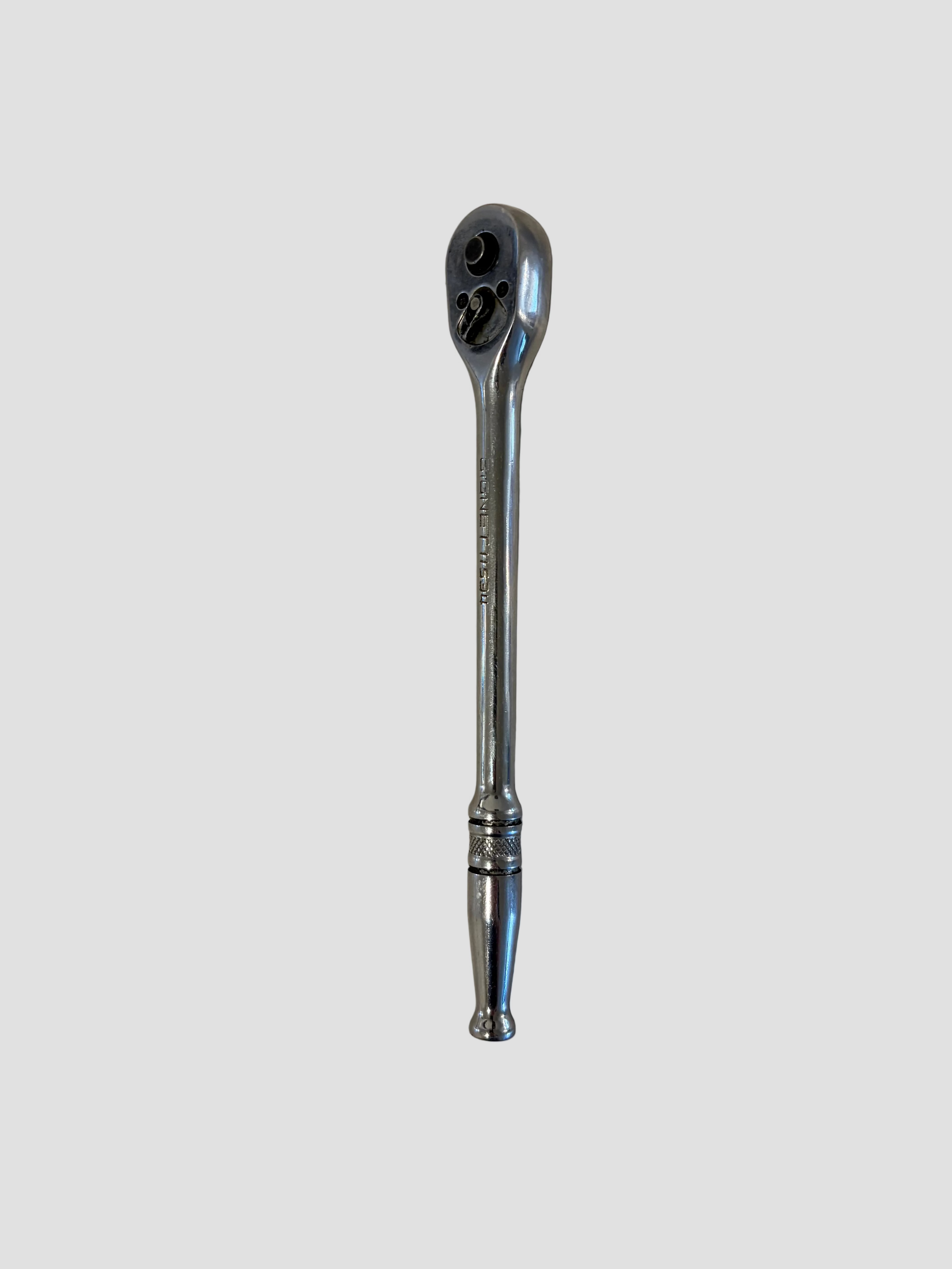 A chrome ratchet wrench lying flat on a light-colored surface. The wrench has a polished metallic finish with a long handle featuring decorative grooves near the end and a rounded ratcheting head containing a square drive socket connector and directional switch. The reflective surface highlights its smooth contours and durable construction, typical of professional-grade hand tools.