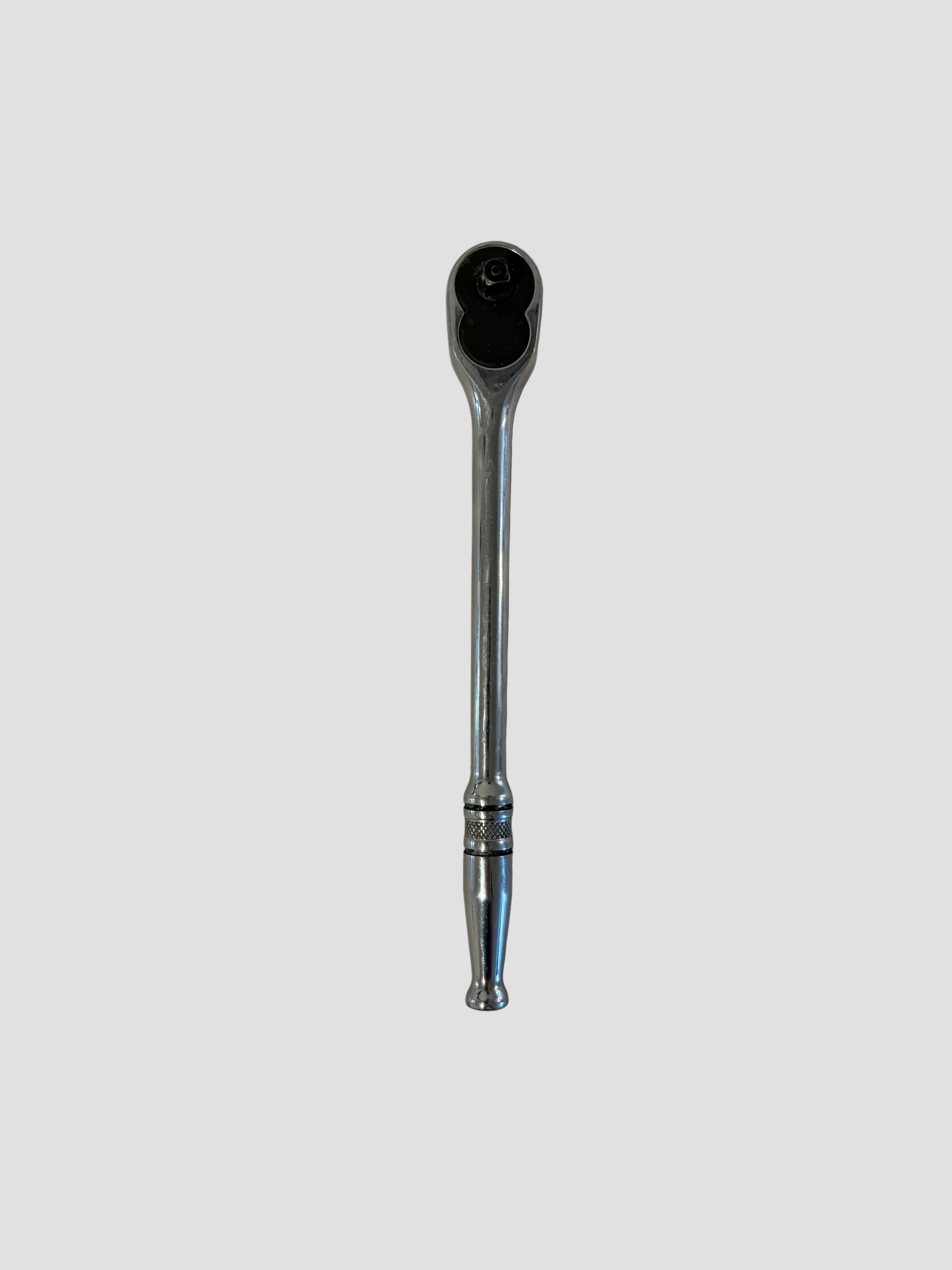 A chrome ratchet wrench lying flat on a light-colored surface. The wrench has a polished metallic finish with a long handle featuring decorative grooves near the end and a rounded ratcheting head containing a square drive socket connector and directional switch. The reflective surface highlights its smooth contours and durable construction, typical of professional-grade hand tools.