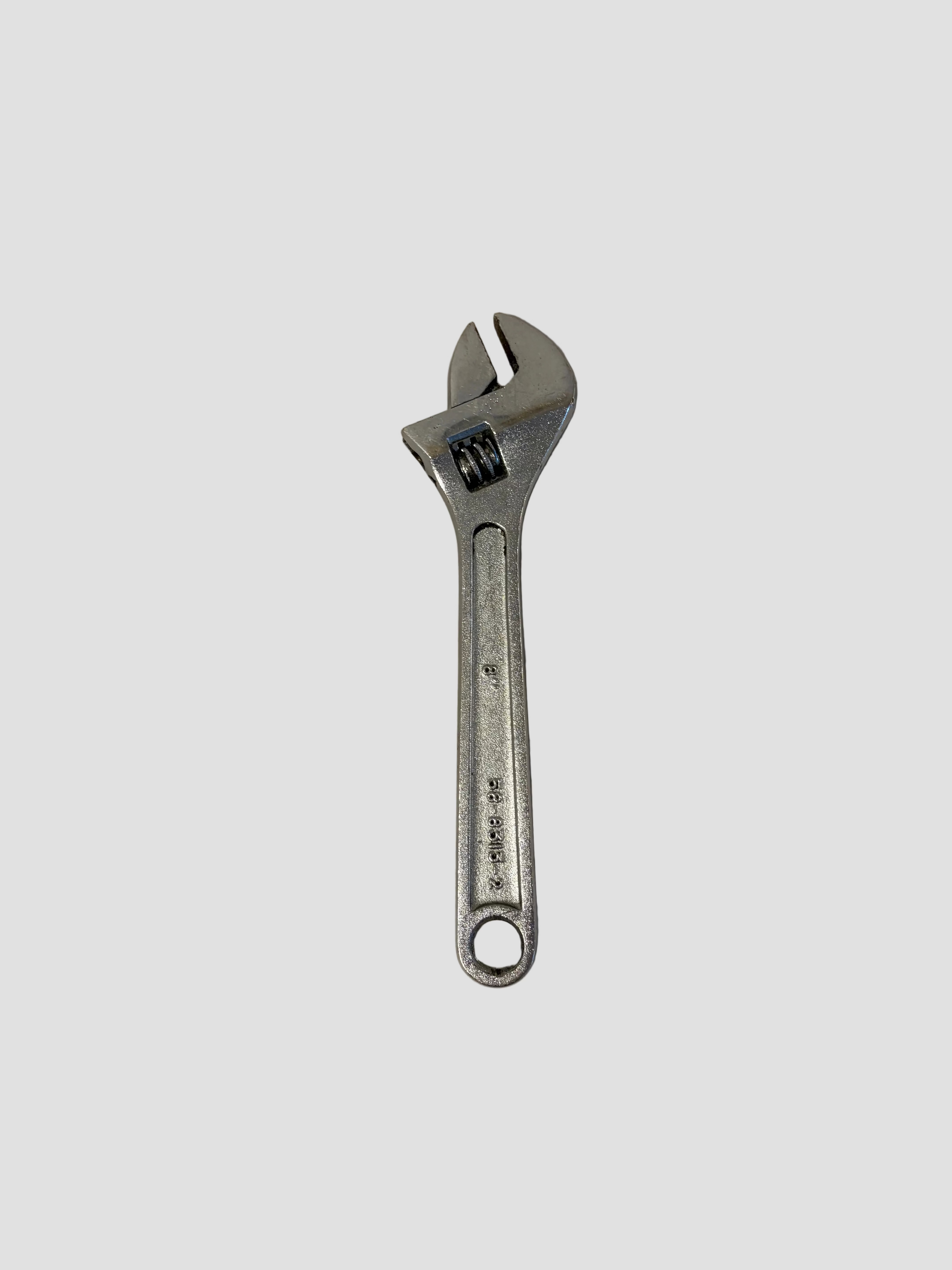 A small silver adjustable wrench resting on a light-colored surface. The wrench has a movable jaw controlled by a worm gear mechanism near the head, allowing it to fit various bolt sizes. The handle features an engraved marking that reads “DROP FORGED,” and a circular hole at the end for hanging or storage. The tool’s polished metal finish and compact design make it suitable for precision mechanical or household tasks.