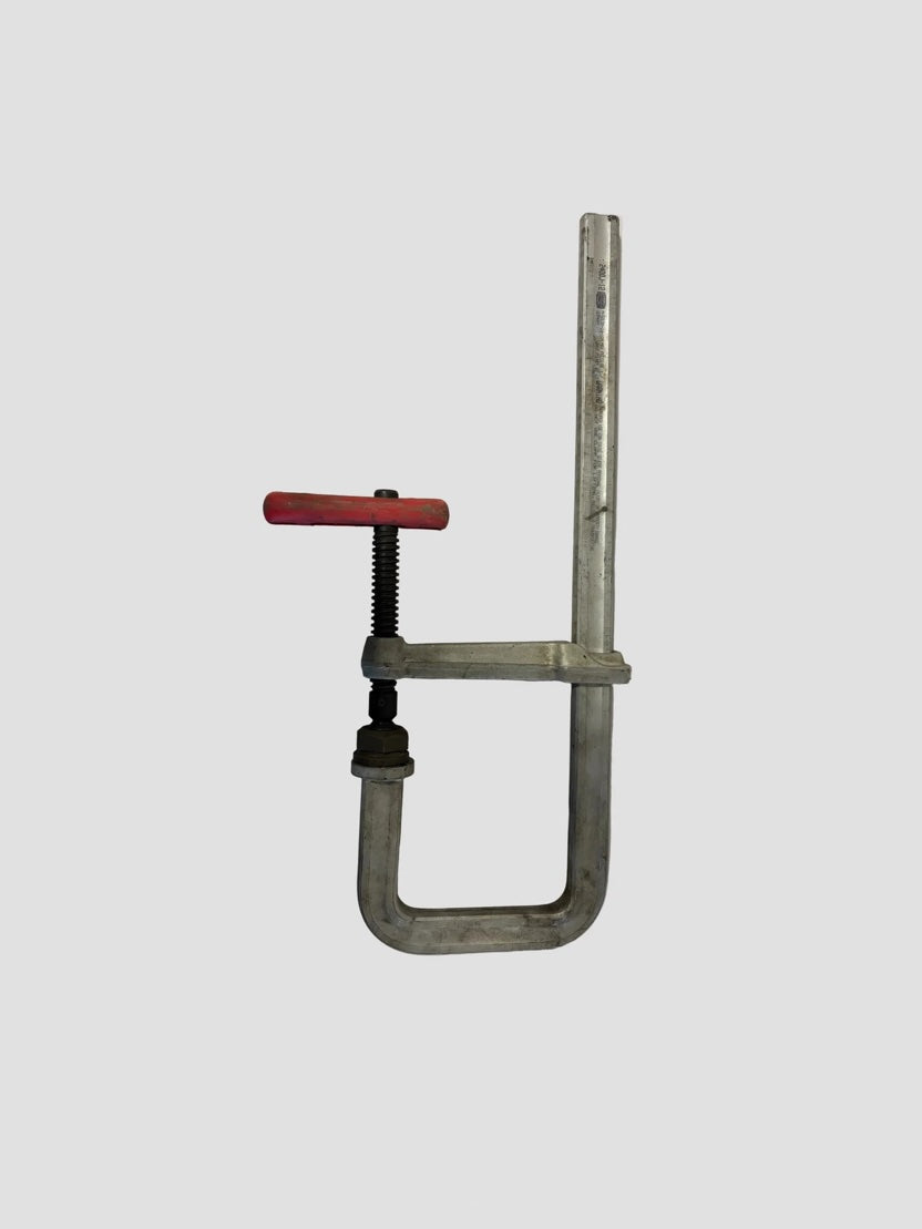 A metal F-clamp with a red handle resting on a light-colored surface. The clamp features a long steel bar with a sliding arm and a fixed arm forming an “F” shape. The red handle is attached to a threaded screw mechanism used to tighten or loosen the clamp’s grip. The metal surfaces show slight wear, indicating regular use, and the brand “BESSEY ST200/2” is stamped on the vertical bar.