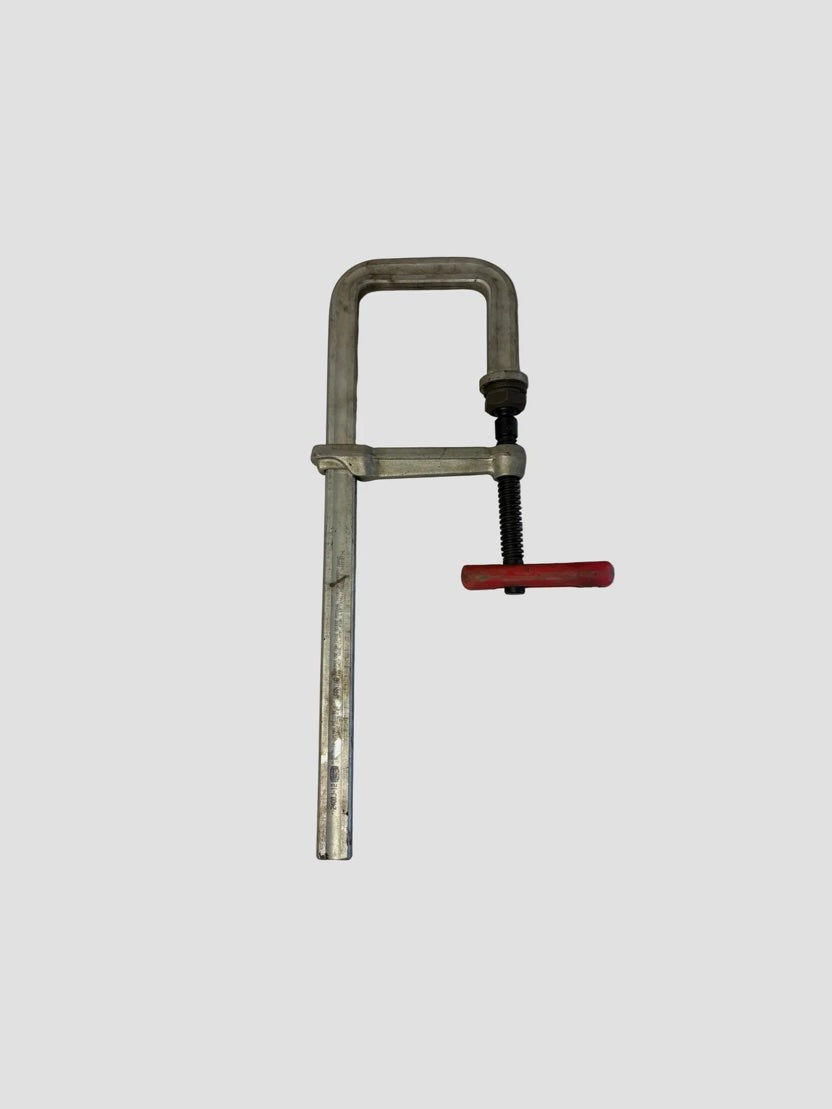 A large metal F-clamp with a red handle resting on a light-colored surface. The clamp features a long steel bar with a fixed jaw at the top and a sliding adjustable jaw below it. The threaded screw mechanism with the red handle allows tightening or loosening the clamp’s grip. The metal surfaces show slight wear and markings, and the brand “BESSEY TG4 100/2” is stamped on the vertical bar, indicating its model and manufacturer.