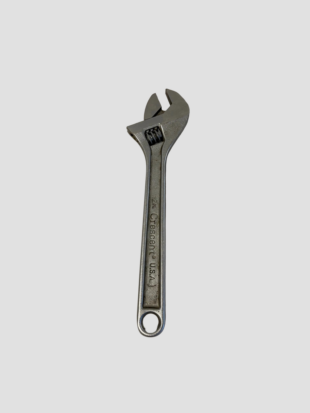 A silver adjustable wrench resting on a light-colored surface. The wrench has a movable jaw controlled by a worm gear mechanism near the head, allowing it to fit various bolt sizes. The handle features an engraved marking that reads “12IN Crescent® U.S.A.,” indicating its size and manufacturer. The tool’s polished metal finish and sturdy design reflect its use for mechanical and maintenance tasks.