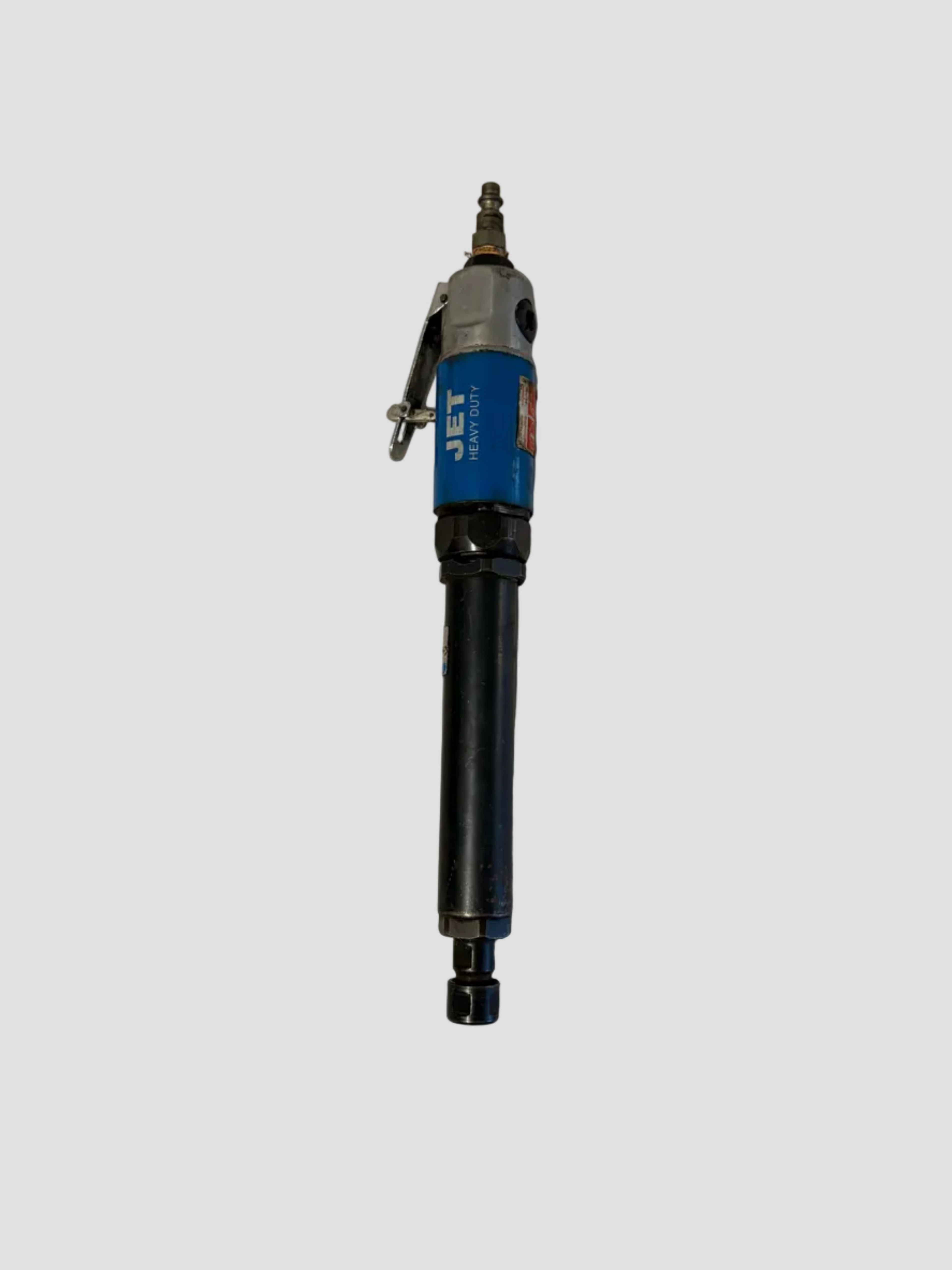 A pneumatic straight air die grinder resting on a light-colored surface. The tool has a cylindrical body with a blue and silver upper section labeled “JET HEAVY DUTY” and a black lower section ending in a collet for attaching grinding bits. A metal lever on the side serves as the trigger, and an air hose connector is attached at the top. The tool’s industrial design and sturdy construction indicate it’s used for grinding, polishing, or cutting metal surfaces in workshop settings.