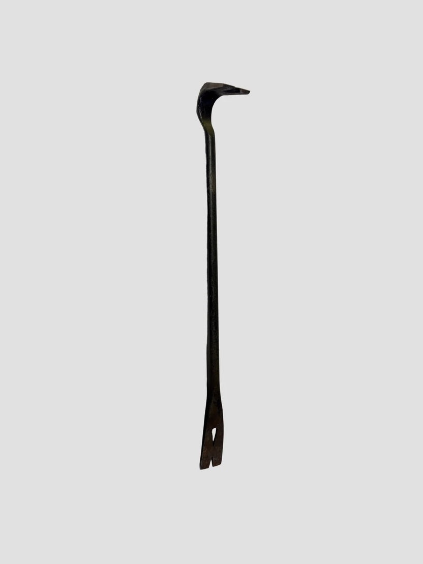 A metal pry bar, also known as a nail puller or crowbar, resting vertically on a light-colored surface. The tool has a dark metallic finish with a curved claw at one end for prying and a flat, forked tip at the other for removing nails. Its sturdy, elongated design shows signs of wear, indicating frequent use in construction or demolition tasks.