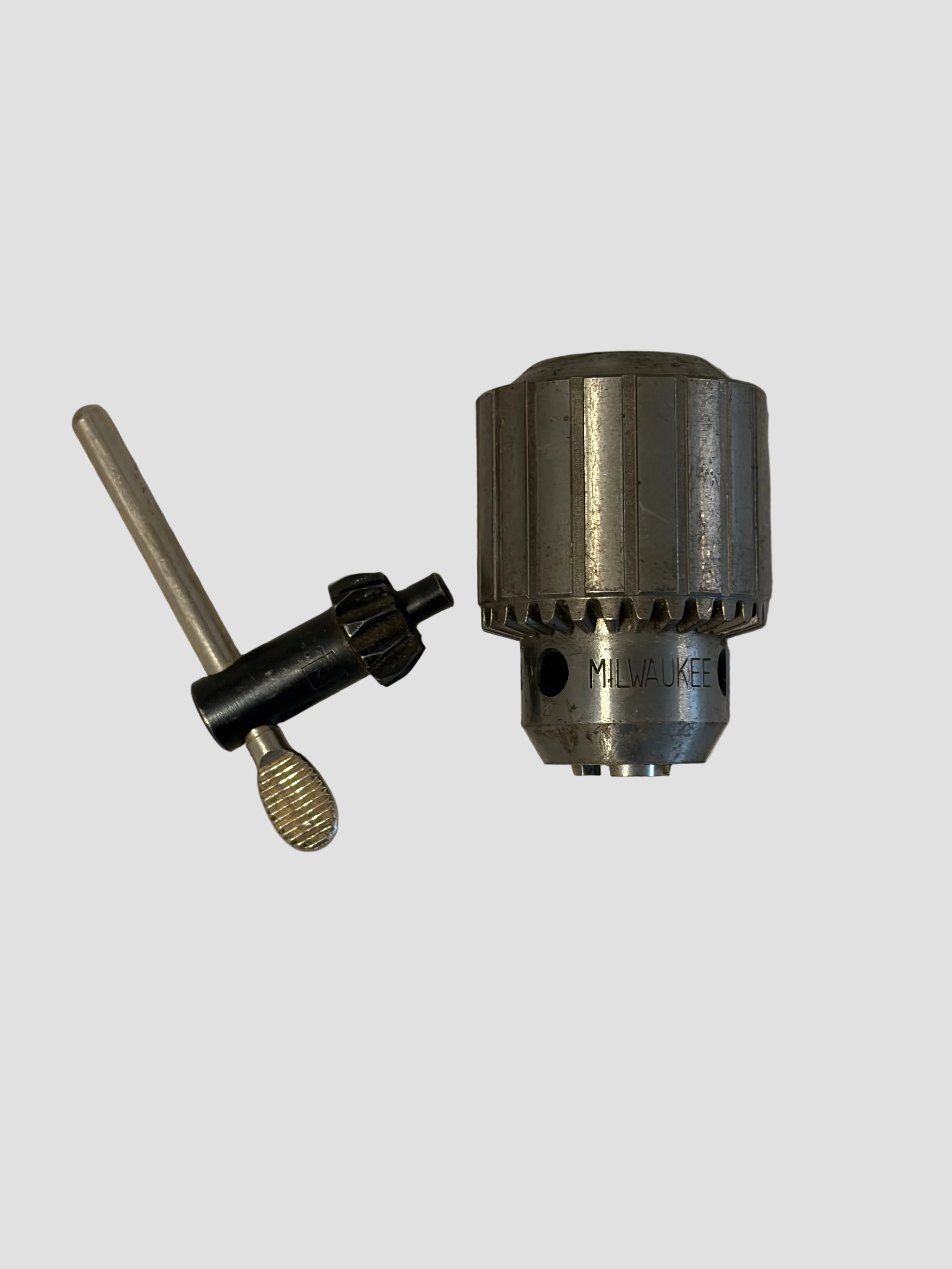 A metal drill chuck with a black and silver chuck key tool, photographed on a light off-white textured surface.