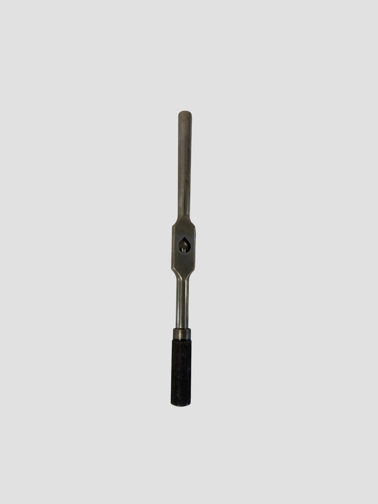 A metal tap wrench positioned vertically against a plain light gray background. The tool features a long cylindrical handle with a central rectangular section containing a square opening for holding a tap, and a textured black grip at the bottom for secure handling. The metallic surface has a matte finish, highlighting its sturdy construction and utilitarian design.